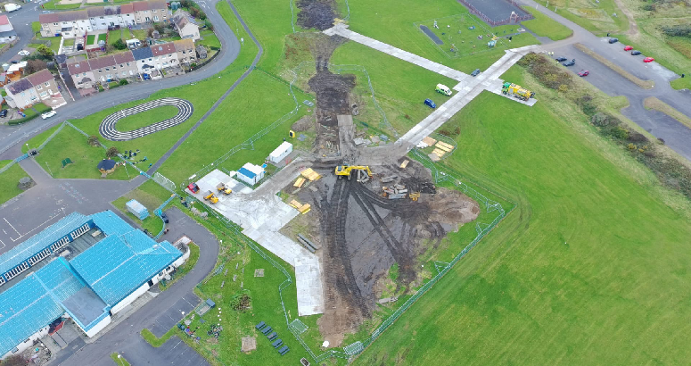 Aerial view of a construction site with heavy machinery and materials, adjacent to a residential area and a car park.