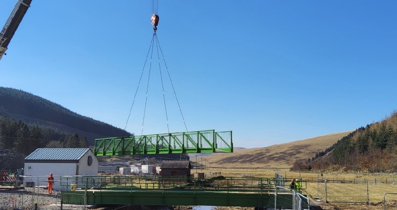 A large green bridge section being hoisted by a crane over a construction site, with workers observing near a small building, set against a backdrop of hills under a clear blue sky.