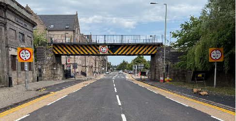 View from the middle of Tay Street road. Buildings line the pathway on the left and trees on the right. A railway bridge can be seen crossing over the road.