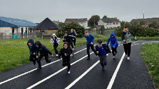 Children running along a running track. Grass surrounds the track and buildings can be seen in the background. The sky is grey.
