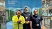 Field Response Clyde Team Celebrates Long-Service Three employees standing in front of a Scottish Water banner inside an office building. The person on the left is wearing a high-visibility vest, and the other two are in casual work attire, all smiling at the camera.