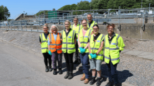 A group of people standing facing the camera wearing hi-vis vests and other PPE. They are standing outside in on a concrete road surface.