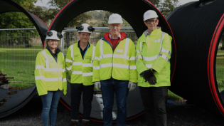 A group of four people wearing PPE including hi-vis, safety goggles, hard hats and gloves, stand outside in front of pipe sections of 2.2 metre diameter. In the background is a grassed area.