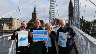 A group of four people stand outdoors on a bridge. They are holding blue and white posters. There are building in the background and a cloudy sky.